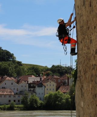 Ein Mitarbeiter mit Helm und Kletterausrüstung beim Seilklettern an einer senkrechten Wand in urbaner Umgebung.