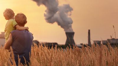 Person mit Kind in einem Feld aus hohem Gras vor einem Industriekomplex mit rauchenden Schornsteinen bei Sonnenuntergang.