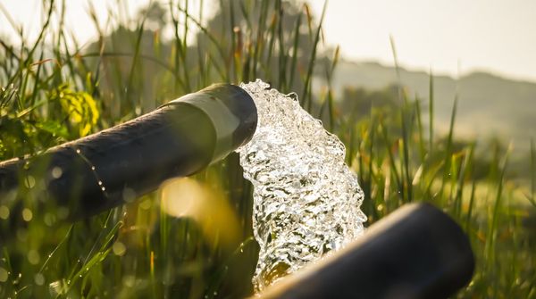 Zwei schwarze Rohre, aus einem fließt klares Wasser in eine grasbewachsene Umgebung im Hintergrund.