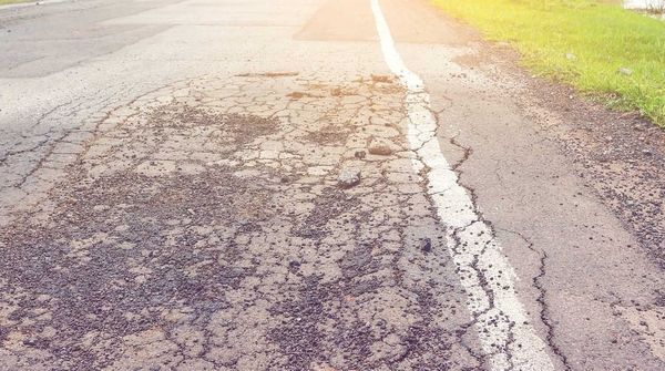 Eine Straße mit Rissen und Kies, im Hintergrund Sonnenlicht, Wiesen und Bäume