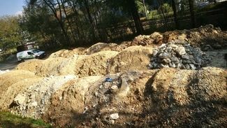 Mehrere große Haufen aus Erde und Bauschutt neben einem Waldstück, im Hintergrund ein Fahrzeug und Gebäude.