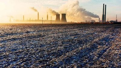 Industrielandschaft mit rauchenden Schornsteinen und Kühltürmen bei Sonnenauf- oder -untergang, umgeben von einem Feld.