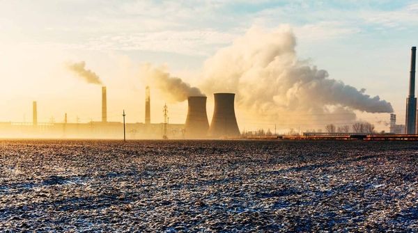 Industrielandschaft mit rauchenden Schornsteinen und Kühltürmen bei Sonnenauf- oder -untergang, umgeben von einem Feld.