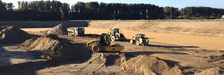 Mehrere Baumaschinen wie Lader und Muldenkipper bewegen und ebnen Sand auf einer offenen Baustelle, umgeben von Bäumen.