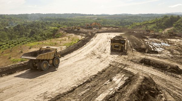 Zwei große Laster fahren auf einem Baustellengelände in ländlicher Umgebung mit grünen Hügeln im Hintergrund.