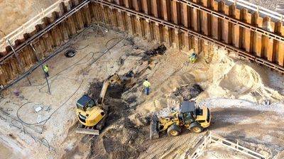Luftaufnahme einer Baugrube, umgeben von Stützmauern, in der mehrere Menschen mit Baustellenfahrzeugen arbeiten.