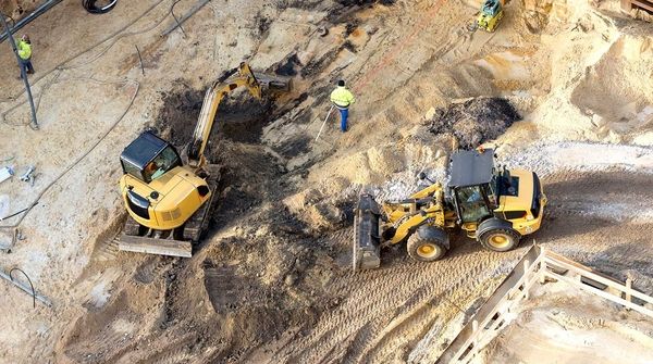 Luftaufnahme einer Baugrube, umgeben von Stützmauern, in der mehrere Menschen mit Baustellenfahrzeugen arbeiten.