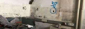 Verschmutzte Innenwand mit Rohren, Steckdosen und blauer Graffiti-Markierung ‚38‘, umgeben von Werkzeugen und Materialien.