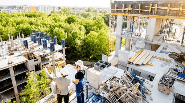 Zwei Männer mit einem Plan in der Hand stehen auf einem Gebäude auf einer Baustelle und schauen auf weitere Arbeiter und Baustellen hinunter.
