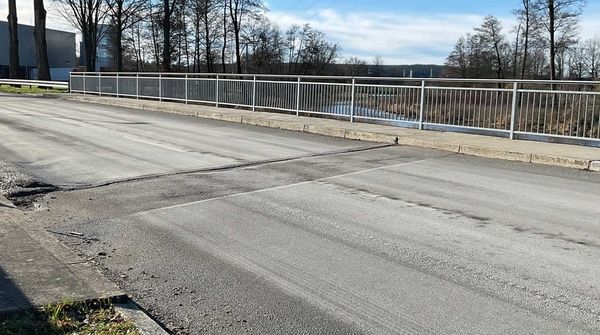 Eine frisch aussehende Straßenoberfläche führt über die Brücke, daneben ein silbernes Geländer und Bäume im Hintergrund.