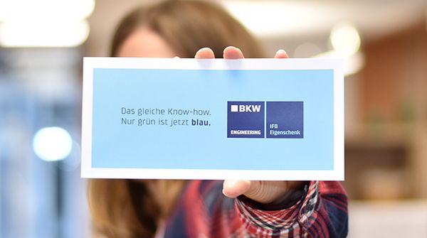 Eine Person hält eine blaue Karte mit dem Text „Das gleiche Know-how. Nur grün ist jetzt blau.“ sowie den Logos von BKW Engineering, IFB Eigenschenk.