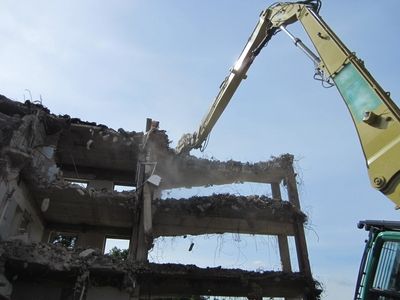Teilweise abgerissenes Gebäude mit drei Etagen wird von einem Bagger mit Hydraulikhammer abgetragen, Staub und Trümmer fallen während des Rückbaus